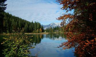 A tranquil lake surrounded by green forests and colorful autumn leaves. In the background, the mountains and a blue sky are visible.