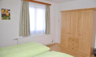 A cozy bedroom with two beds and bedding in a green checkered pattern. Windows with curtains and a wood-colored wardrobe complete the room.