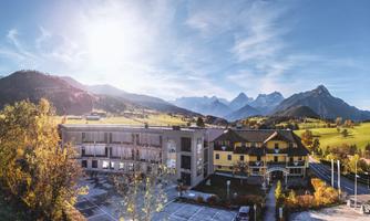A picturesque landscape with mountains in the background and two buildings in the foreground. The sun shines brightly, creating an inviting atmosphere.