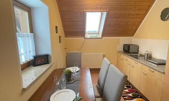 A cozy kitchen with a dining area and a wooden table. The walls are light, and there is a window opening with natural light.