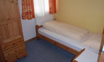 A simple bedroom with a single bed, a dresser, and a wardrobe. The walls are yellow and there is a large window with red curtains.