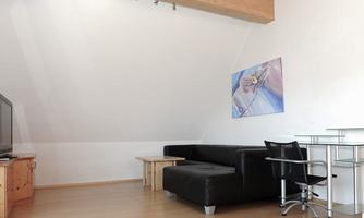 A minimalist living space with a black sofa and a small table. A modern picture hangs on the wall, and there is a TV as well as a glass table with a chair.