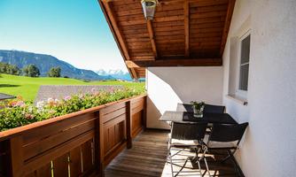 A cozy balcony with a table and chairs. In the background, green meadows and mountains can be seen.