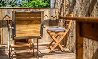 A cozy wooden balcony with a folding chair and another chair. The surroundings are green and invite relaxation.