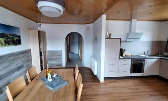 A modern kitchen with a large wooden dining table. The room is bright and friendly with wooden ceilings and a picture on the wall.