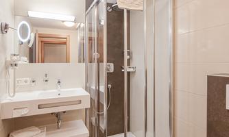 A modern bathroom with a shower and a sink. The walls are light-tiled and there are large mirrors.