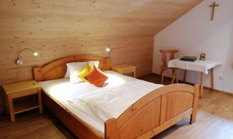 A cozy bedroom with a wooden bed and a soft pillow. There are nightstands and a small table in the corner, as well as a cross on the wall.