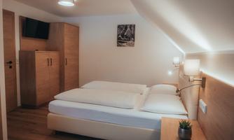 A cozy bedroom with a double bed and modern wooden interior. The walls are painted light and a small plant stands on the bedside table.