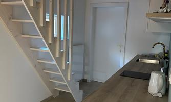 A bright living room with a modern staircase and a small kitchenette. The walls are painted in neutral colors and the atmosphere is friendly and inviting.