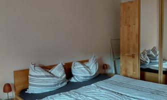 A cozy bedroom with a large bed and decorative pillows. There is a wooden wardrobe and a bedside table that complement the room.