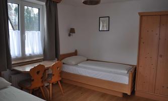 A simple room with two single beds, a table, and chairs. Large windows let in plenty of daylight.