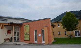 A modern building with an orange facade and large windows stands in front of a green meadow. In the background, mountains and a tree are visible.