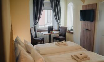 A cozy hotel room with two single beds and a window. There is a small table with chairs and a television on the wall.