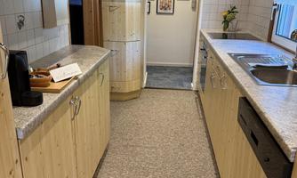 A modern kitchen with wooden furniture and a long countertop. Pictures hang on the wall, and the floor is carpeted.