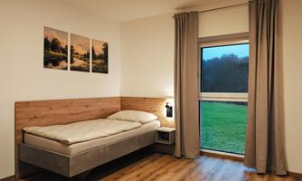 A simple bedroom with a single bed, a nightstand, and a window overlooking a green meadow. On the wall, there are two pictures of a landscape.