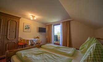 A cozy room with two beds and checked pillows. There is a small table, a television, and plenty of daylight coming through the window.