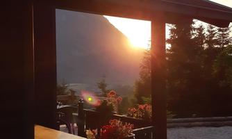 A wooden building overlooking mountains and plants in the foreground. The sun is setting behind the mountains, creating a beautiful atmosphere.