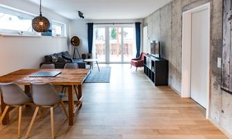 A modern living room with a large table and comfortable chairs. Large windows provide plenty of natural light and access to the outdoor area.