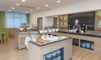A modern dining hall with a self-service buffet and a chef preparing dishes. The decor is bright and friendly, with wood details and large windows.