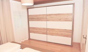 A modern bedroom with a large wardrobe in wood and white tones. The room is bright and friendly, with a warm wooden floor and soft lighting.