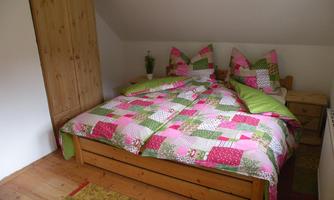 A cozy bedroom with a large bed and colorful bedding. A wooden wardrobe stands against one wall, and the floor is covered with a carpet in various colors.