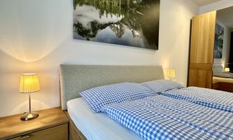 A cozy bedroom with a large bed and checked bedding. A landscape painting hangs on the wall, and there is a bedside lamp.