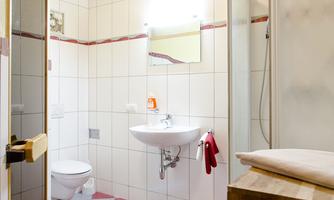 A modern bathroom with white tiles and a red and white checkered pattern on the floor. There is a shower, a sink, and a small wooden shelf.