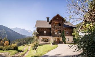 A charming mountain house surrounded by green meadows and mountains in the background. The sky is clear and the atmosphere is calm and inviting.