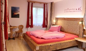 A cozy bedroom with a large wooden bed and red pillows. The walls are bright, and there is a small table as well as a window with curtains.
