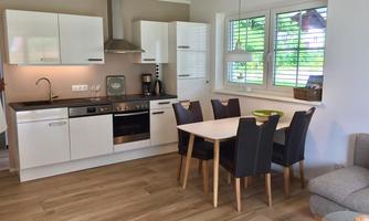 A modern kitchen with light furniture and a dining table for six people. Large windows provide plenty of daylight and a view of the outdoors.