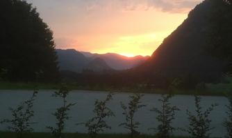 A picturesque sunset behind the mountains, accompanied by gentle hills. The scene is framed by trees and a green meadow.