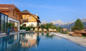 A modern outdoor pool surrounded by comfortable loungers and a rustic wooden house. In the background, majestic mountains are visible under a clear blue sky.