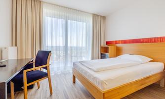 A modern hotel room with a double bed, a desk, and a chair. Large windows provide plenty of daylight and offer a view outside.