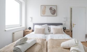 A bright bedroom with a double bed and decorative pillows. The walls are white and there is a piece of art above the bed.