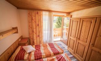 A cozy bedroom with two beds and wooden paneling. Large windows offer a view of the balcony and nature.
