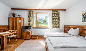 A cozy bedroom with two beds and wooden furniture. Large windows provide plenty of daylight and a view of the landscape.