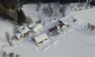 A tranquil snowy landscape with several buildings surrounded by deep snow. In the background, snow-covered trees can be seen.