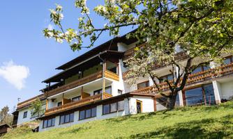 A modern house with several balconies, surrounded by green meadows and blooming trees. The sky is clear and blue, creating a cheerful atmosphere.