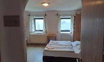 A cozy bedroom with a double bed and wooden floor. There are windows with curtains and a small table in the room.