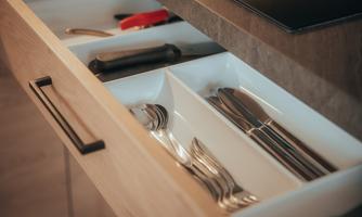 A modern kitchen drawer with cutlery in white inserts. Additionally, some kitchen utensils are visible, such as a peeler and a spatula.