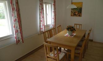 A bright dining room with a large wooden table and chairs. Red-and-white curtains hang at the windows, and there is a vase of flowers on the table.