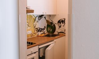 A small modern kitchen with white cabinets and wooden flooring. A decorative mosaic is visible on the wall.