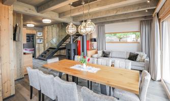 A modern, bright living room with a large dining table and comfortable chairs. The wooden paneling and large windows create a cozy atmosphere.