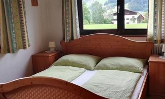 A simple bedroom with a wooden bed and two green pillows. A green landscape is visible through the window.