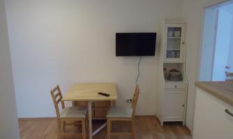 A small dining area with a wooden table and two chairs. A television hangs on the wall, next to which is a cabinet display.