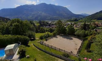 A picturesque landscape with a riding arena and beautiful mountains in the background. In the foreground, gardens and a pool are visible.