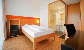 A modern hotel room with a single bed, a desk, and a wardrobe. There is a glass shower in the bathroom area and wooden flooring.