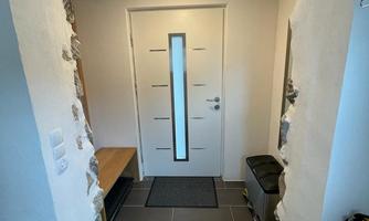 A modern entrance area with a white door and a narrow window. The floor is tiled with dark tiles, and there is a bench and a trash can in the room.