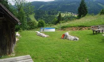 A green meadow with stones and a wooden terrace. In the background, mountains and a swimming pool can be seen.