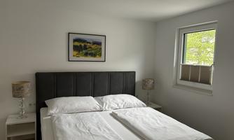 A bright bedroom with a double bed and two nightstands. A painting hangs on the wall and a window lets in plenty of daylight.